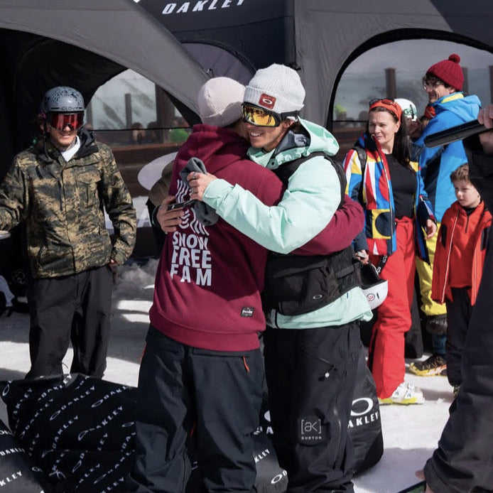 Two snowboarders hugging on a snowy slope with Oakley branding in the background